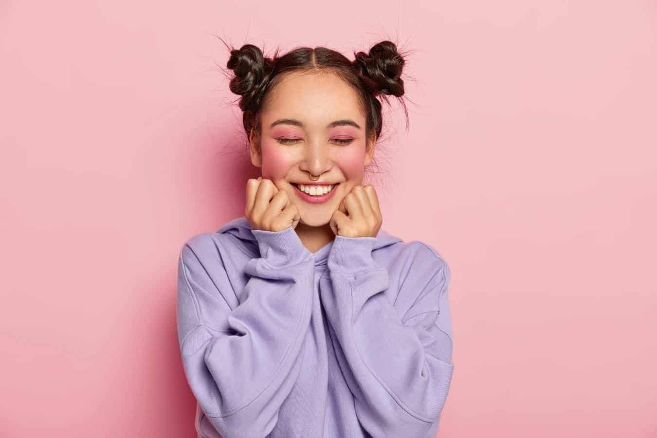 Asian girl with her hair in two buns is smiling and pinching her cheeks against a pink background.