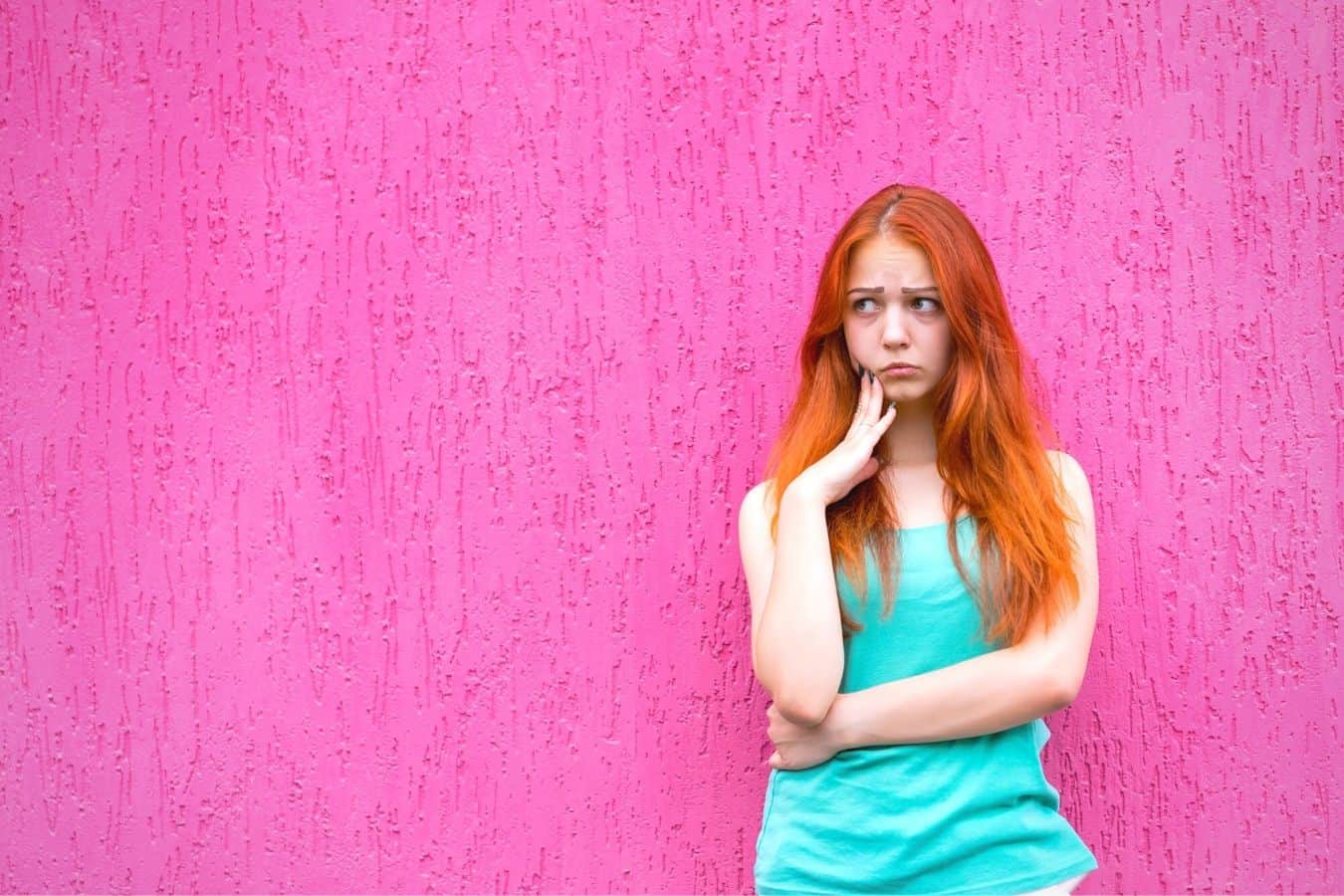 Woman with long red hair looking sad, stood in front of a pink wall.