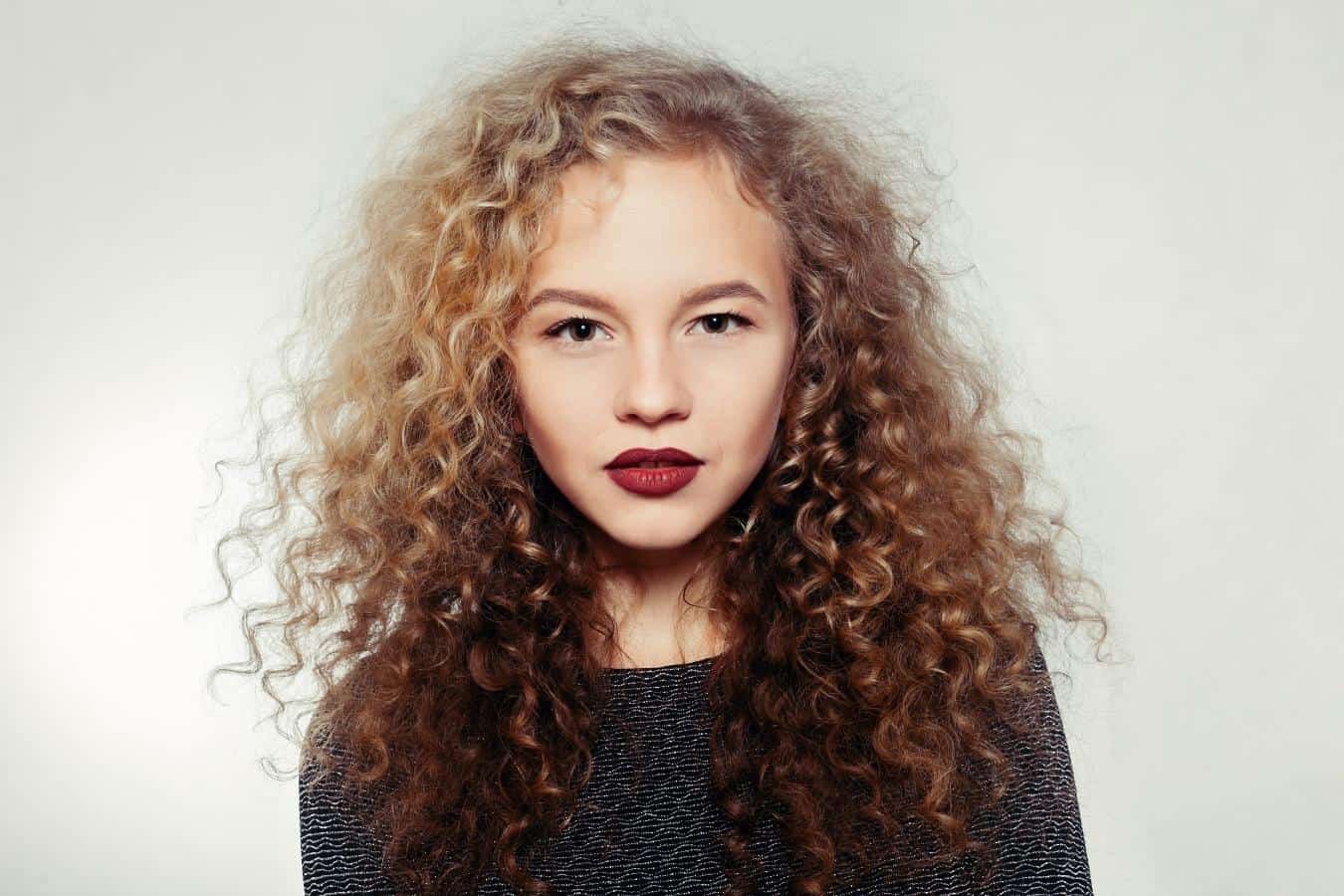 A woman with very curly hair, staring straight at the camera against a white background.