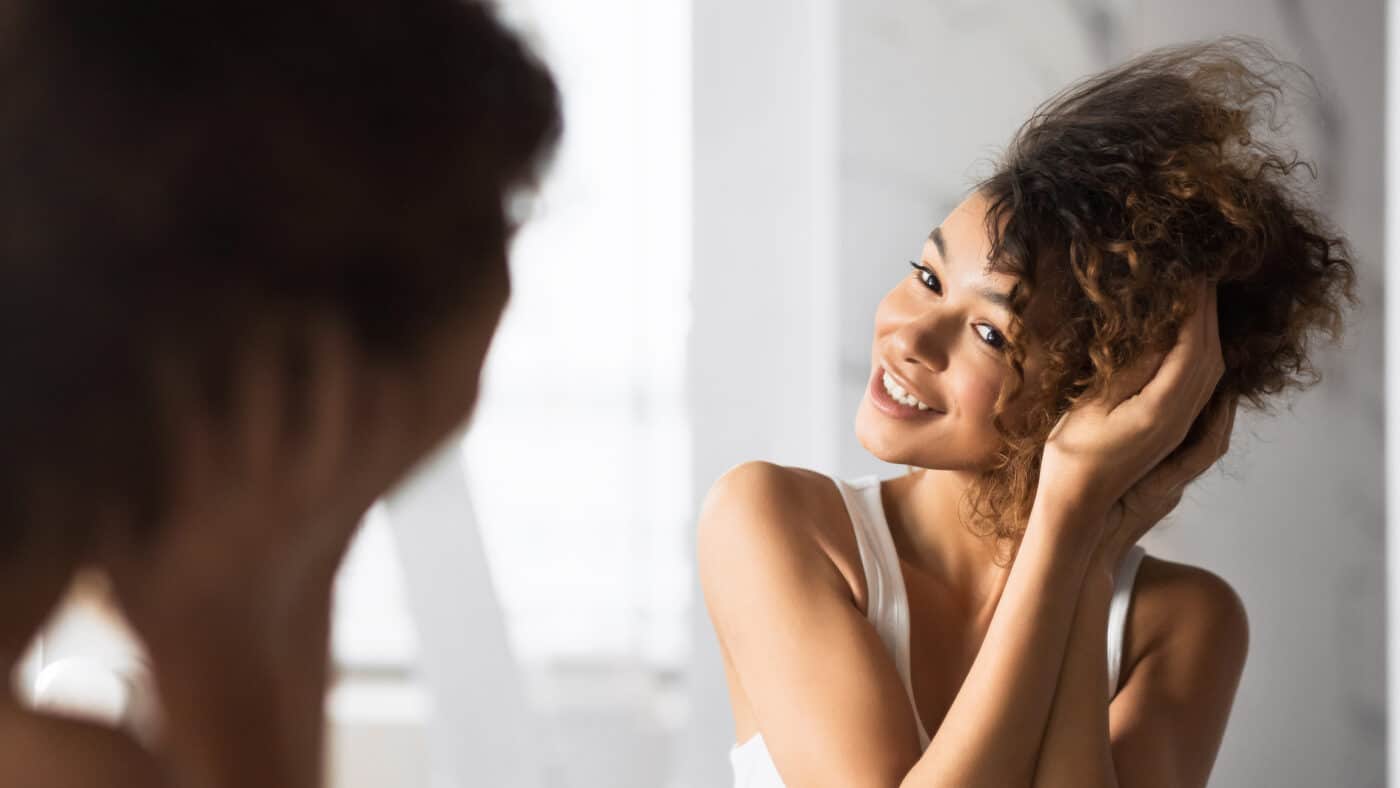 Woman is smiling and looking into the mirror and touching her curly hair at the same time